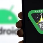 Hand holding a smartphone displaying the Android 15 logo on screen, with the blurred Android mascot logo visible in the background