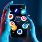 Hands holding a smartphone with floating app icons including Facebook, Twitter, heart, location pin, music, video, email, and browser apps against a blue gradient background with sparkle effects
