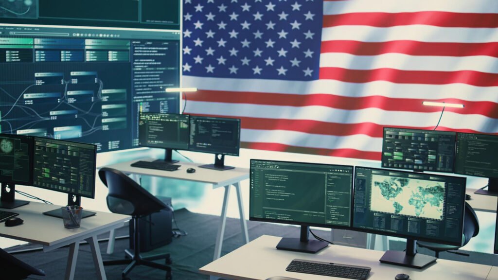 Modern government office workspace with multiple computer monitors displaying software audit dashboards and data analytics, with American flag in background representing US federal agency technology operations